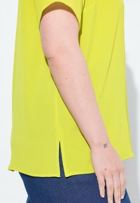 Top a maniche corte, di un vivace colore giallo, realizzato in tessuto strutturato, con fessure laterali all'orlo e una vestibilità ampia, abbinato a jeans blu scuro.