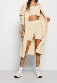 Beige textured lounge set featuring a cropped top and shorts, paired with a long coat, and accented with white sneakers and a scrunchie.