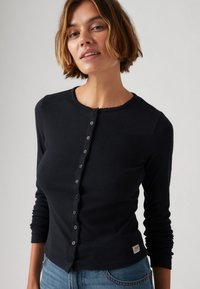 Melnas garās piedurknes pogājamā džemperis ar strukturētu ribu, apaļu kaklu un viļņotu malu; tajā ir mazas melnas pogas un logo apakšmalā.