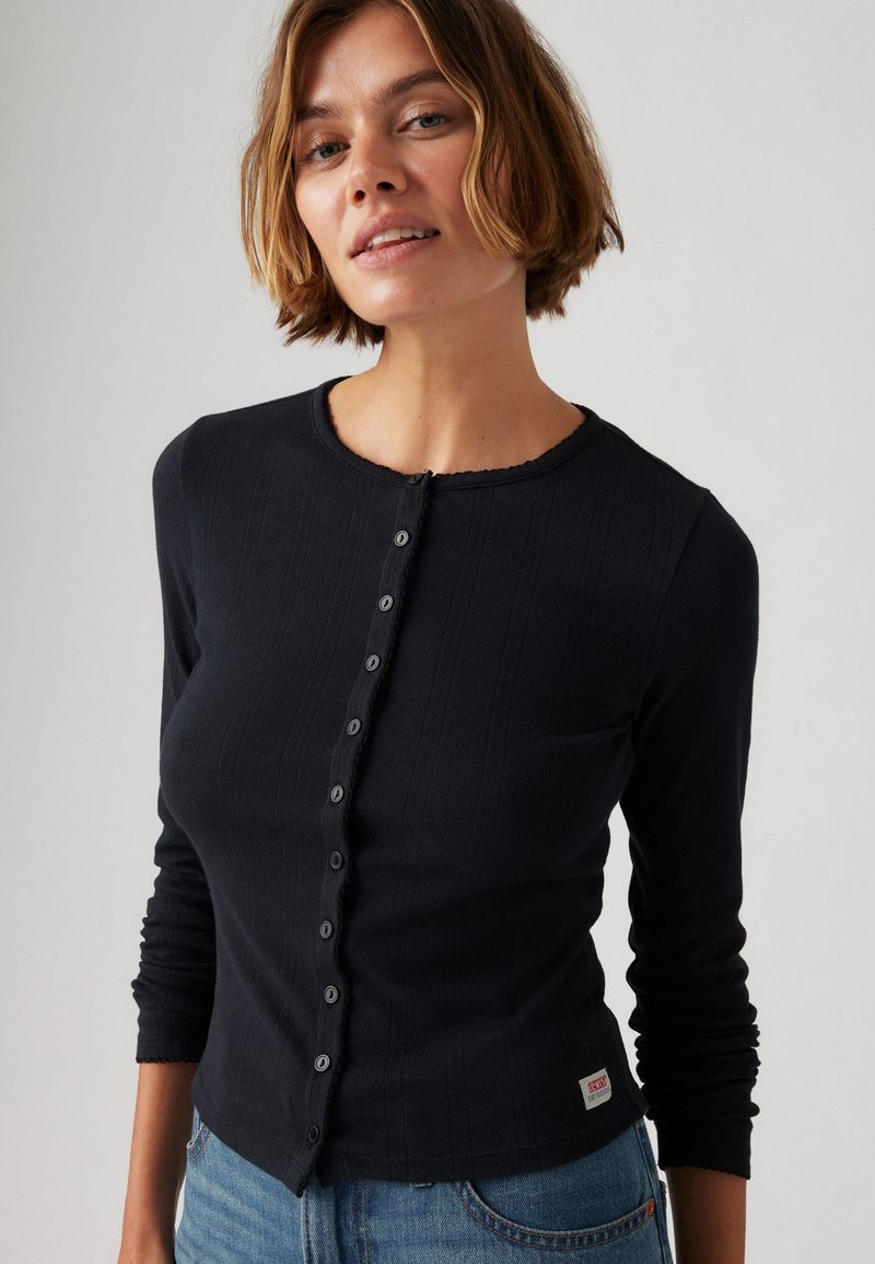 Cardigan noir à manches longues avec boutons, bordures texturées, col rond et bords festonnés ; présente de petits boutons noirs et un logo à l'ourlet.