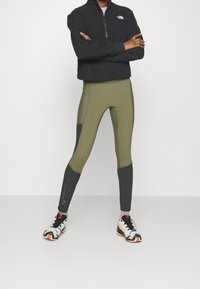 Grüne und graue Sportleggings aus dehnbarem Stoff, mit hohem Bund und seitlichen Einsätzen, kombiniert mit einer schwarzen Fleecejacke mit Reißverschluss und Turnschuhen.
