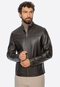 Baardige man met een donkerbruin leren jack over een beige coltrui, met beide handen de rits van het jack vasthoudend.