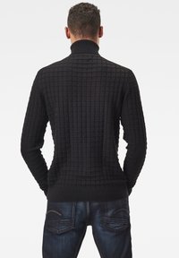 Svart strukturerad tröja med hög krage och rutmönster, med långa ärmar och ribbade ärmslut som smalnar av mot handlederna.