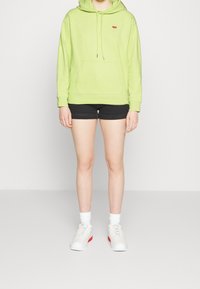 Sudadera de algodón verde lima con un bolsillo frontal y cordones, combinada con pantalones cortos de mezclilla negros y zapatillas blancas con acentos rojos.