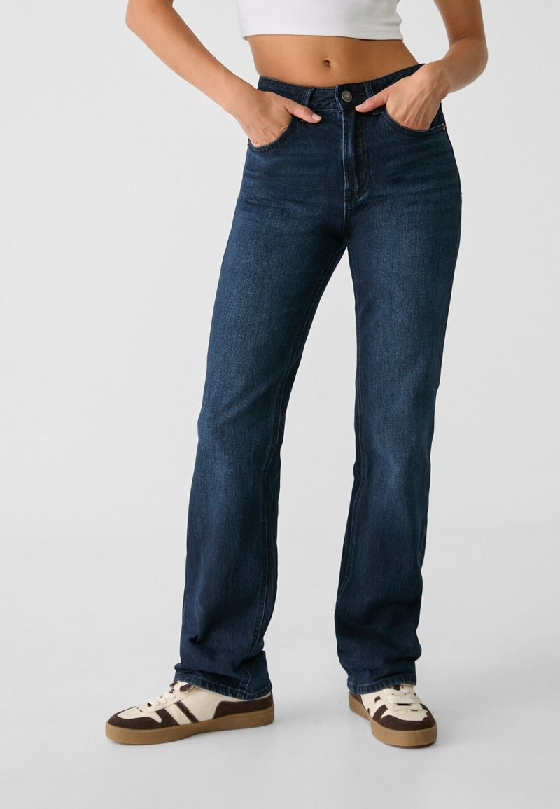 Personne portant un jean droit bleu foncé, un crop top blanc et des baskets blanches avec des détails marron, les mains dans les poches avant.