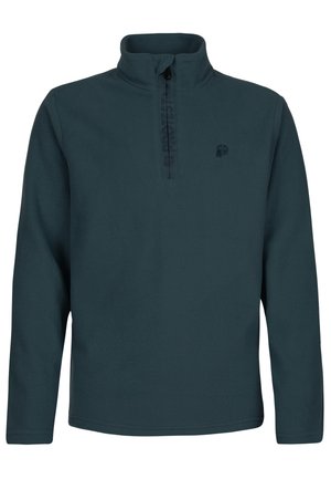 Jongere met een marineblauwe muts, donker vest met rits, blauwe sneeuwbroek, zwarte laarzen en zwarte zonnebril, staand met handen in de zakken.