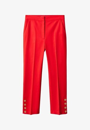 Personne porte un pull en maille blanche côtelée avec une fermeture éclair à l'avant et un pantalon rouge sur mesure, les mains dans les poches, debout devant un fond uni.