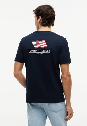 Mann trägt ein marineblaues Tommy Hilfiger T-Shirt mit rotem, weißem und blauem Logo und dem Schriftzug "Tommy Hilfiger Since 1985" auf der Rückseite.