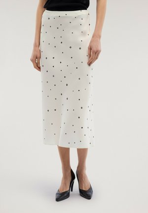 Femme portant une jupe midi blanche à pois noirs, associée à des chaussures à talons noirs et un haut noir, debout devant un fond uni.