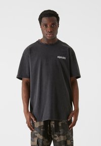 Sort oversized t-shirt lavet af bomuld, med korte ærmer og rund halsudskæring, prydet med små hvide bogstaver trykt på brystet.