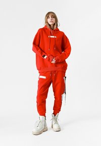 Sudadera roja con cordones y detalles en blanco, combinada con pantalones de chándal rojos a juego que tienen bolsillos laterales y detalles bordados. Botas blancas.