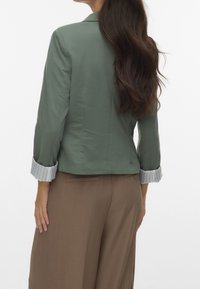 Blazer vert au design structuré, avec des manches rayées retroussées. Associé à un pantalon brun à jambes larges, mettant en valeur des détails subtils.