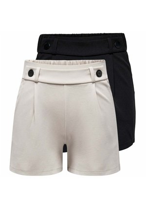 Junge Frau steht, trägt ein weißes kurzärmliges Oberteil mit schwarzem Blumendesign, schwarze Shorts und schwarze Ballerinas vor einfachem Hintergrund.