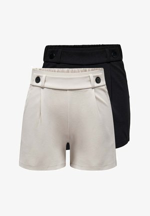 Junge Frau steht, trägt ein weißes kurzärmliges Oberteil mit schwarzem Blumendesign, schwarze Shorts und schwarze Ballerinas vor einfachem Hintergrund.