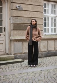 Hnědý fleecový svetr na zip s oversized rukávy, černé kalhoty s širokými nohavicemi a tmavě šedý šátek. Oblečení doplňují neformální boty.