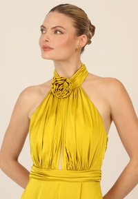 Gelbes Trägerkleid aus glattem Stoff, mit Falten, einem floralen Rosettendetail am Ausschnitt und einem Schlüsselkernausschnitt.