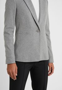 Une personne porte un blazer gris clair ajusté avec un seul bouton sur un pantalon noir sur mesure, sur un fond blanc uni.