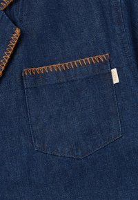Denim fabric in dark blue with orange contrasting stitching. Features a square patch pocket with a label at the side. Smooth texture.