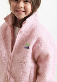 Chaqueta de forro polar rosa con una textura suave. Presenta un cuello alto, cremallera a juego y una insignia en la parte frontal.