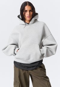 Una donna con i capelli bagnati indossa una felpa oversize grigio chiaro, una maglietta scura a strati e pantaloni cargo verdi oliva, con le mani nelle tasche della felpa.