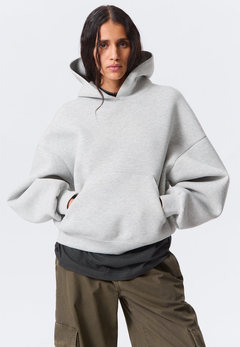 Una donna con i capelli bagnati indossa una felpa oversize grigio chiaro, una maglietta scura a strati e pantaloni cargo verdi oliva, con le mani nelle tasche della felpa.