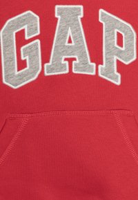 Punainen huppari, jossa on harmaa brodeerattu "GAP"-logo, etupuolella kengurutasku ja teksturoitu kangas. Pehmeä puuvillaseosmateriaali.