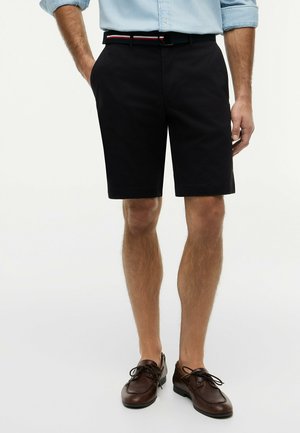 Mann trägt schwarze Shorts, hellblaues hochgekrempeltes Hemd und braune Leder-Schnürschuhe, steht mit den Händen in den Taschen.