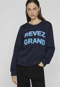 Jūros pajėgų džemperis su didelėmis, tekstūruotomis mėlynomis "REVEZ GRAND" raidėmis, suapvalintu kaklu ir šonkaulinėmis rankovėmis, derinamas su tamsiomis kelnėmis.