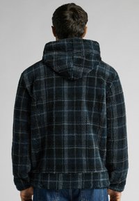 Fleece mikina tmavě modré barvy s kostkovaným vzorem, která má jemnou texturu a kapuci. Design zahrnuje rovný lem a manžety na rukávech.