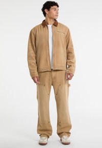 Giacca di jeans beige con colletto in corduroy marrone, abbinata a pantaloni larghi coordinati con grandi tasche e dettagli di cucitura in vista.