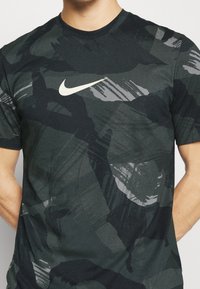 Mörkgrön Nike-t-shirt med ett kamouflagemönster i grå nyanser, rund hals, korta ärmar och en framträdande vit logotyp på bröstet.