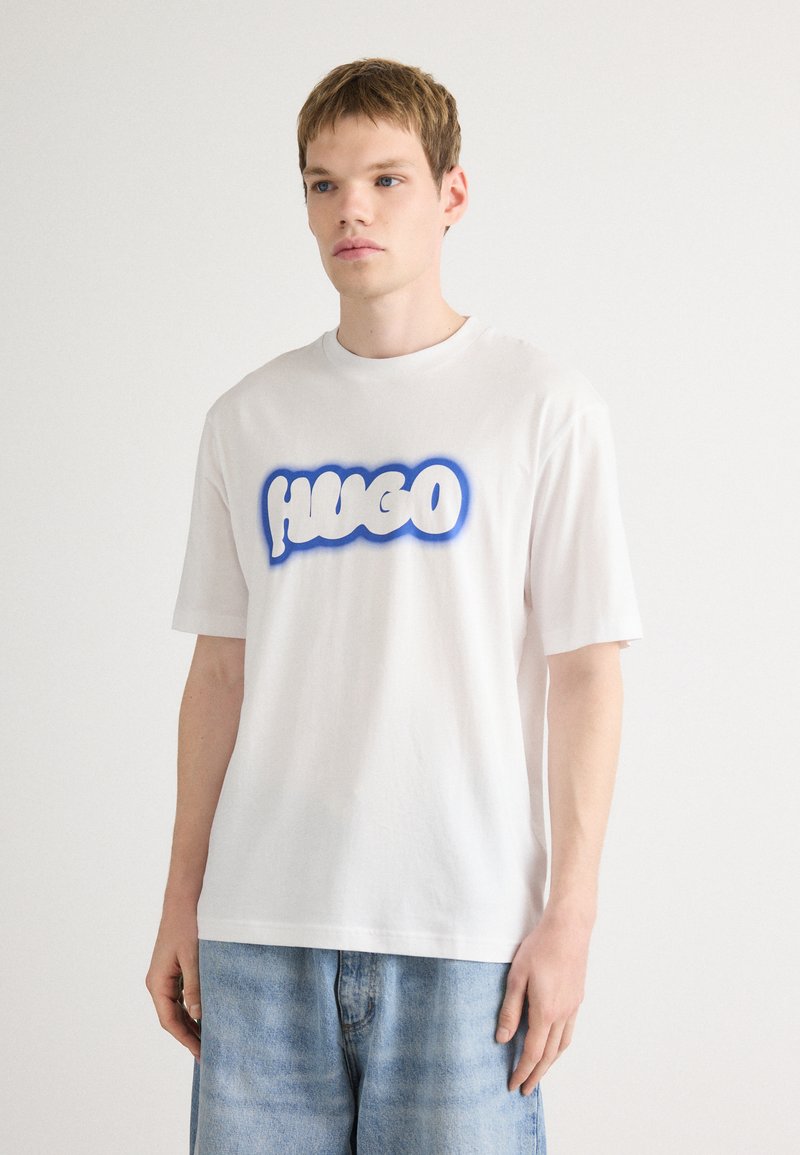 Hvid bomulds-t-shirt med et stort blåt grafisk logo "HUGO" på forsiden, korte ærmer og en afslappet pasform.
