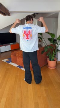Biały bawełniany t-shirt z grafiką homara i logo „ADIDAS” na plecach, zestawiony z czarnymi spodniami o szerokich nogawkach. Wnętrze z roślinami.