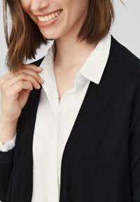 Czarny kardigan na białej koszuli z kołnierzykiem. Kardigan ma miękką, dzianinową teksturę i otwarty fason.