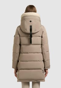 Persona che indossa un cappotto invernale imbottito beige con un ampio cappuccio foderato in pile color crema, visto da dietro.