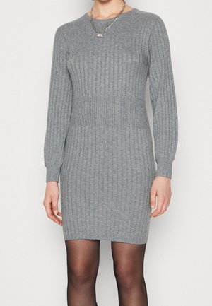 Femme portant une robe gris ajustée en maille côtelée à manches longues, des collants noirs transparents et un collier chaîne argentée.