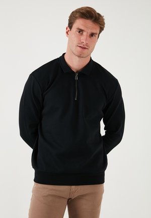 Black pullover with a zip collar, made of a textured fabric. Long sleeves and fitted waist. Worn with light brown pants.