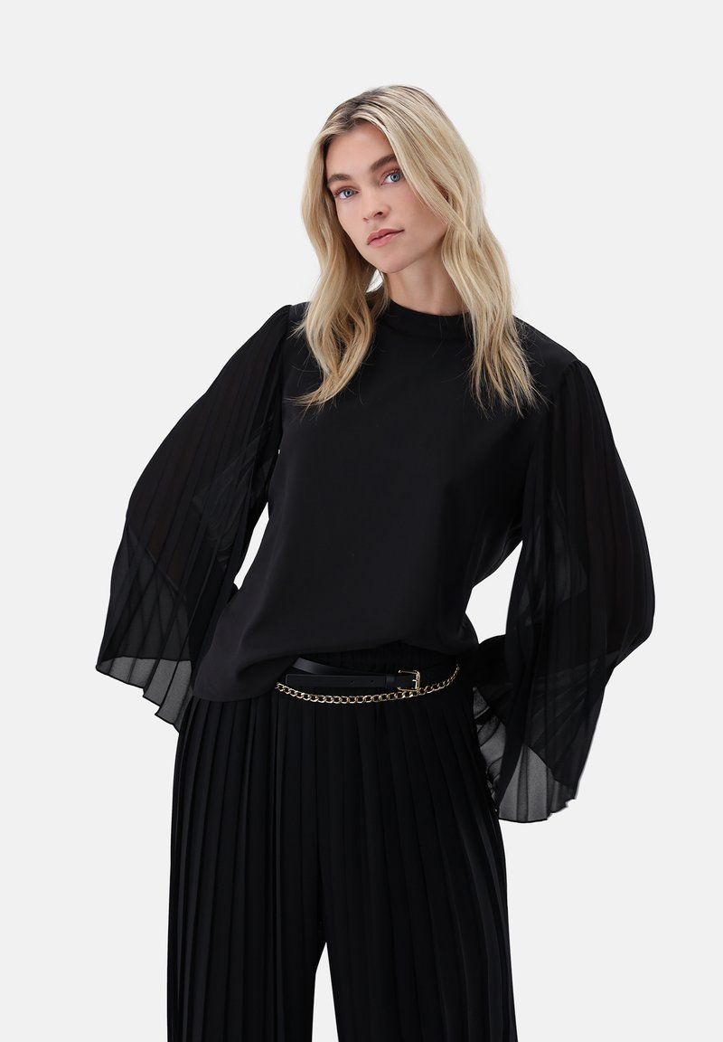 Junge Frau mit blondem Haar trägt eine schwarze Bluse mit transparenten, plissierten Ballonärmeln und schwarze, plissierte Hosen mit einem goldenen Kettengürtel.