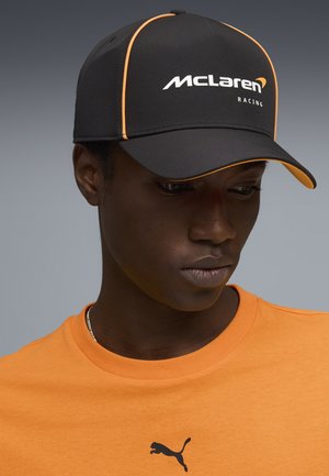 Giovane uomo che indossa un cappellino nero McLaren Racing con rifiniture arancioni e una t-shirt arancione Puma, guarda verso il basso su uno sfondo grigio.