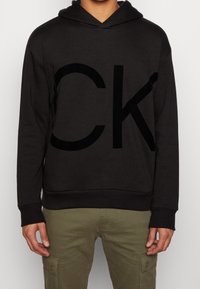 Sudadera con capucha negra con un gran logo "CK" en negro. Hecha de tela suave con un ajuste relajado y un bolsillo tipo canguro. Cuenta con puños y dobladillo acanalados.