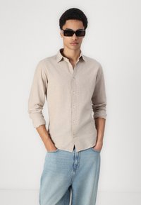 Beige button-up shirt in lightweight fabric with long sleeves. Paired with light blue baggy jeans. Model wearing dark square sunglasses.