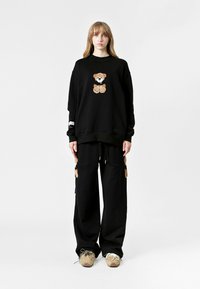 Conjunto de sudadera y pantalones negros que presenta un diseño de oso de peluche bordado. Los pantalones cuentan con detalles de oso en los laterales y un ajuste relajado.
