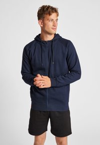 Felpa con zip di colore blu navy, dotata di cappuccio regolabile e maniche raglan, realizzata in un tessuto morbido. Abbinata a pantaloni corti neri. Il modello ha le mani unite.