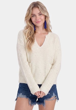 Junge Frau mit langen blonden Haaren trägt einen cremefarbenen Strickpullover mit Pailletten und ausgewaschene Jeansshorts, lächelt sanft.