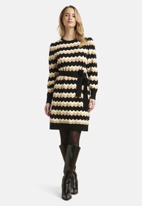 Knie-lange jurk met een zwart, crème en beige zigzagpatroon. Breiselstructuur, lange mouwen en een zwarte ceintuur. Zwarte kniehoge laarzen.