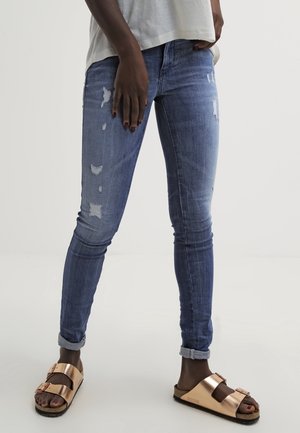 Persona que lleva jeans ajustados azules desgastados con dobladillos enrollados, una camiseta blanca y sandalias metálicas doradas de doble tira, de pie contra un fondo liso.