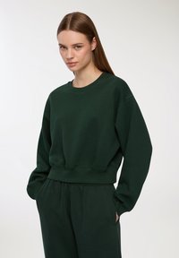 Ciemnozielony sweter z okrągłym dekoltem, wykonany z miękkiego materiału, z opuszczonymi ramionami, ściągaczami oraz luźnym krojem. W zestawie z pasującymi spodniami.
