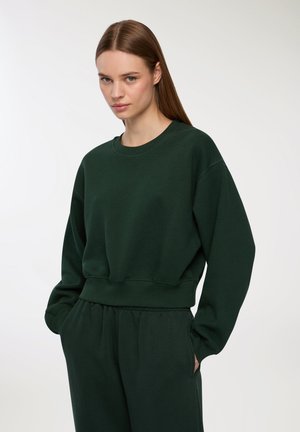 Felpa girocollo verde scuro realizzata in tessuto morbido, con spalle scese, polsini a costine e una vestibilità rilassata. Abbinata a pantaloni coordinati.