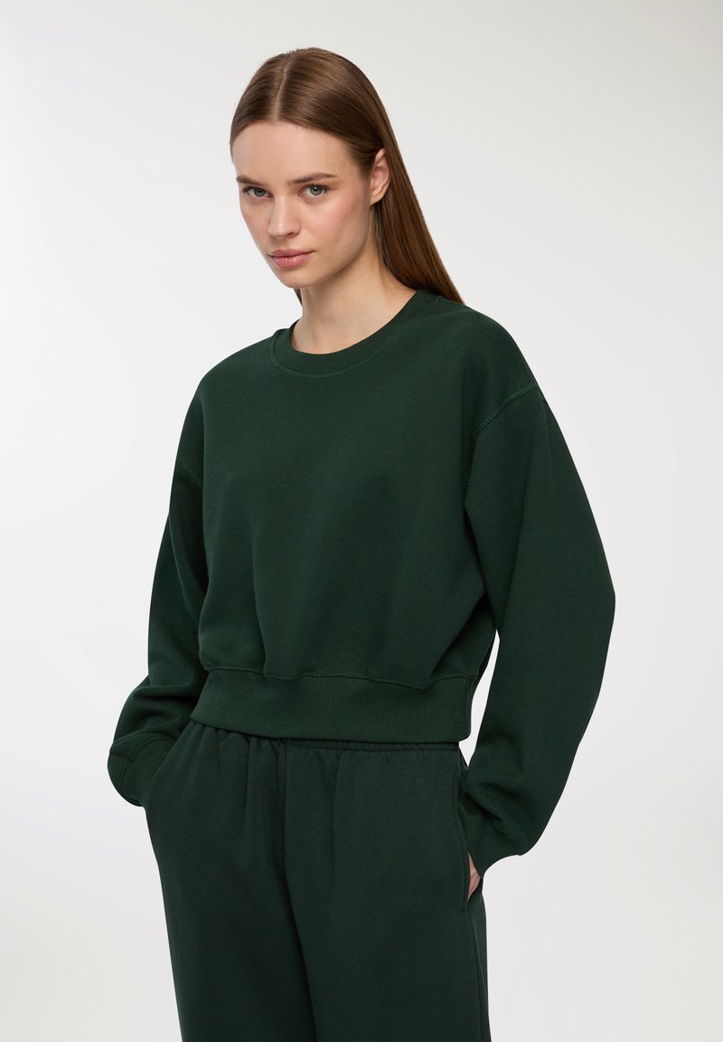 Ciemnozielony sweter z okrągłym dekoltem, wykonany z miękkiego materiału, z opuszczonymi ramionami, ściągaczami oraz luźnym krojem. W zestawie z pasującymi spodniami.