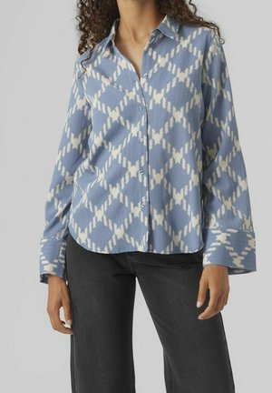 Chemise à manches longues à boutons de couleur bleu clair avec un motif de losanges blancs. Tissu texturé, col pointu et larges poignets. Associée à un jean foncé.